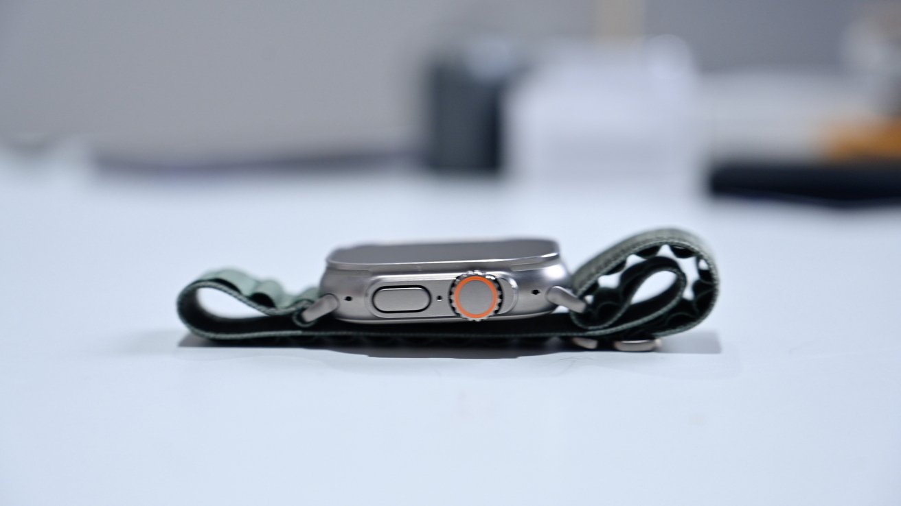 A smartwatch with a side button and orange-rimmed crown lies on a white surface, attached to a grey fabric band.
