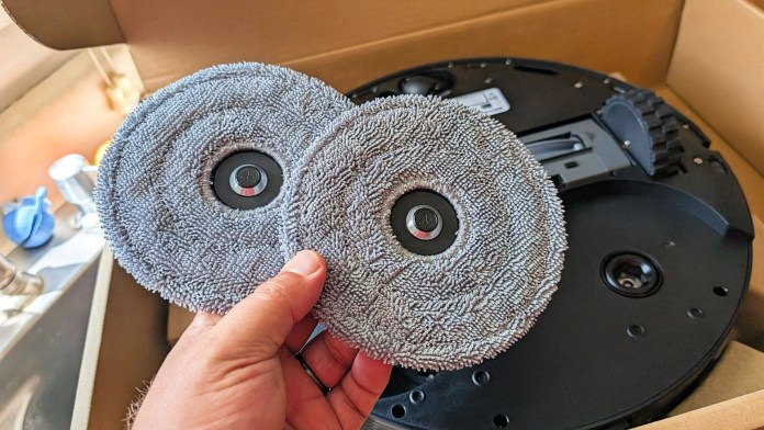 A person holding two microfiber mop pads designed for a robot vacuum, with the robot's base visible in the background.