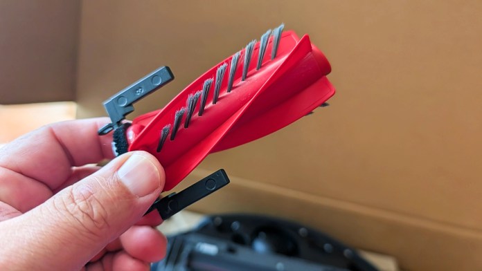 A hand holding a red cleaning tool designed for removing hair and debris from a robot vacuum cleaner's brush, set against a blurred background.