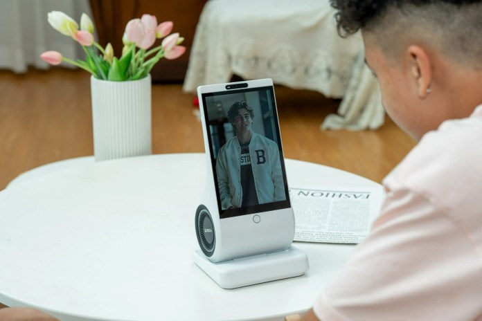 A person interacts with the EUVOLA emotional AI companion device, featuring an 8-inch HD touchscreen displaying an expressive avatar, on a round table surrounded by a vase of flowers.