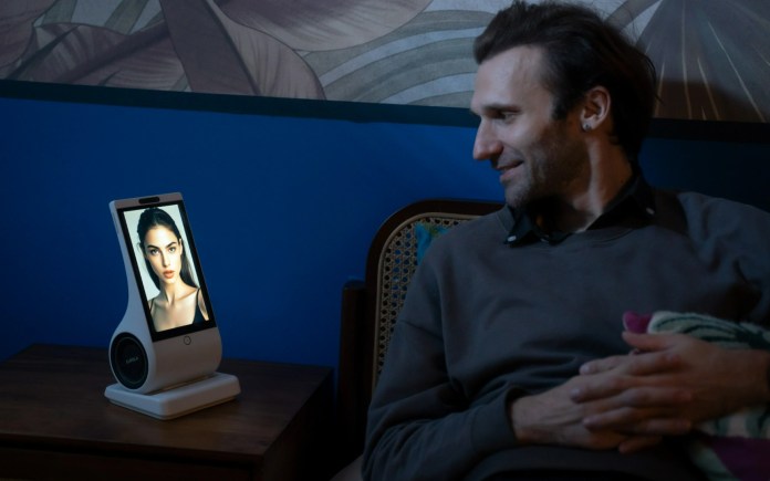 A man sitting beside a table with an EUVOLA emotional AI companion device, which has an 8-inch HD touchscreen displaying a lifelike avatar, set against a dark blue wall with patterned decor.