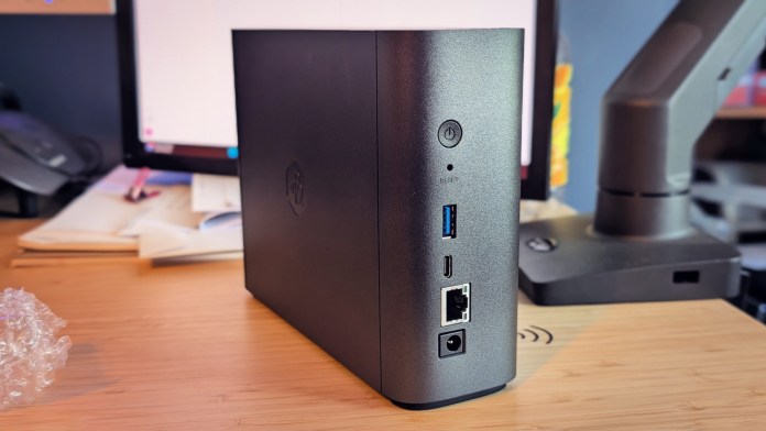 Side view of the Synology BeeStation Plus, showing various ports including USB, Ethernet, and power, with a desk and computer monitor in the background.