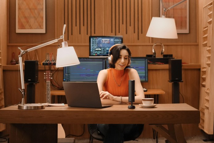 A young woman wearing headphones sits at a wooden desk, smiling while working on a laptop. In front of her is a sleek microphone and a cup of coffee, with audio equipment visible in the background.