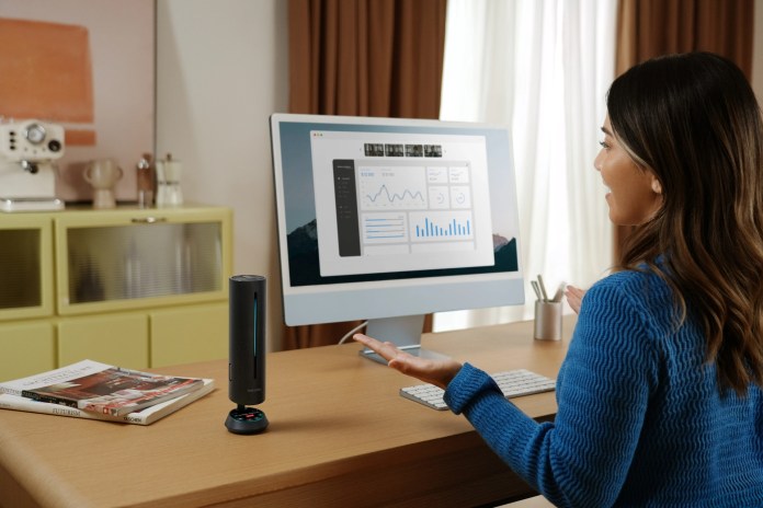 A woman in a blue sweater is engaged in a meeting while using the Insta360 Wave speakerphone, which sits on her desk next to a computer displaying data analytics.