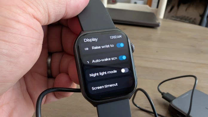 Hand holding a smartwatch displaying settings related to the display, including options for raise wrist to activate, auto-wake screen, night light mode, and screen timeout.