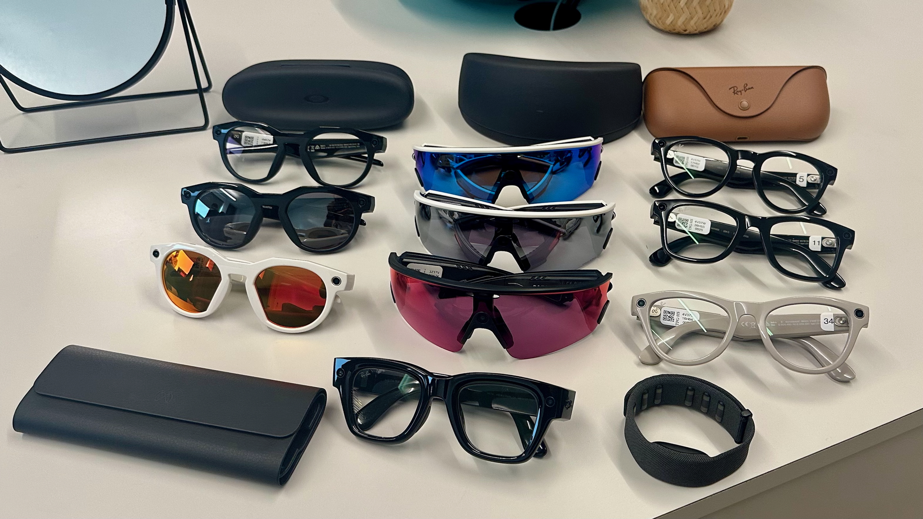 Three Oakley Meta HSTN, three Oakley Meta Vanguard, and three Ray-Ban Meta (Gen 2), along with cases, sitting atop a table in various styles, along with the Meta Ray-Ban Display glasses in front of them all.