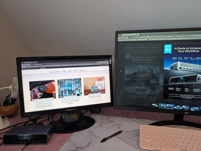 A modern workspace featuring two monitors: the left monitor displays a website about a smartwatch, while the right monitor showcases Anker Prime Docking Station promotional information. Anker's docking station sits prominently on a marble desk, accompanied by a pink keyboard and stationery.