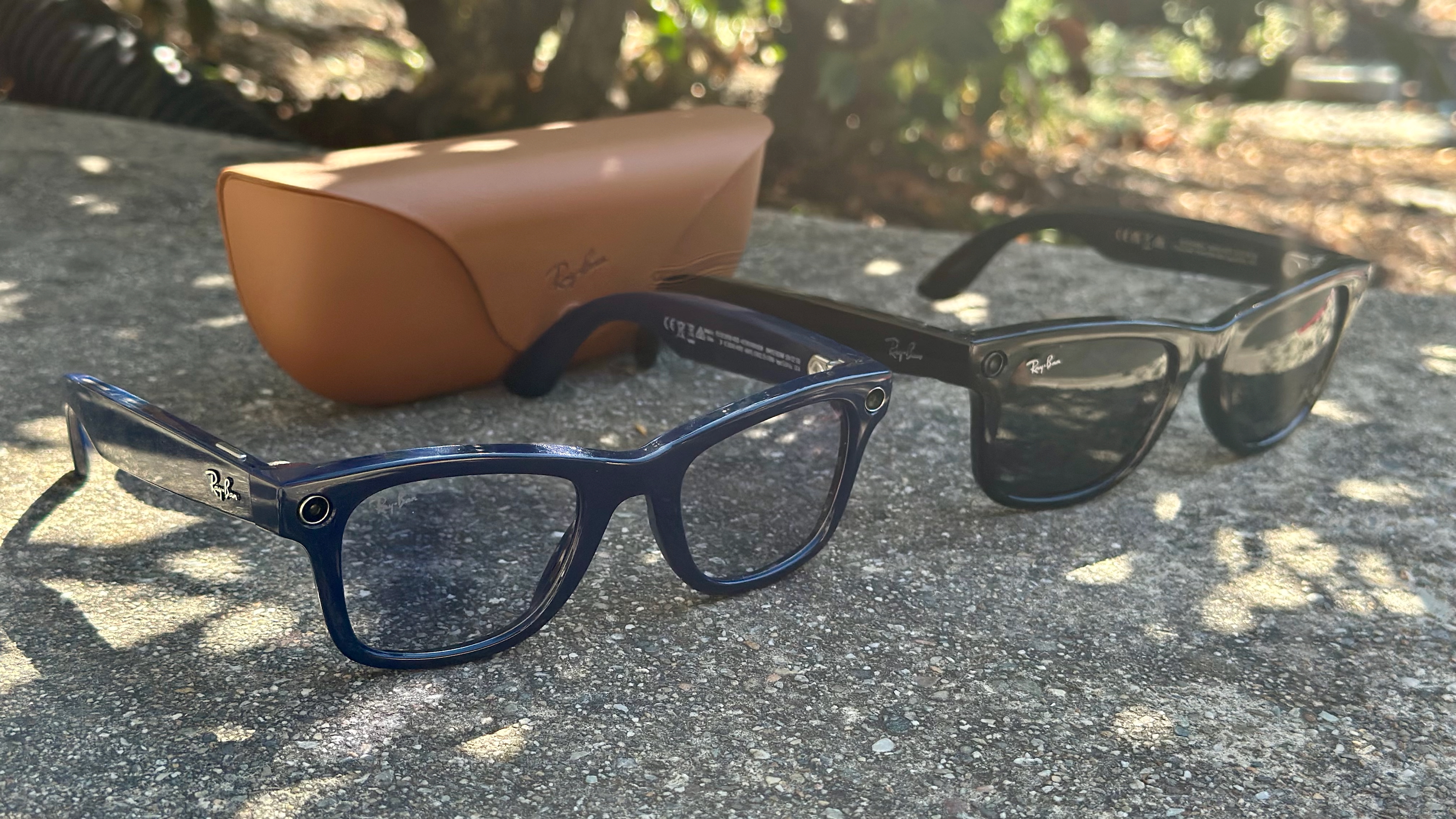 Photo of the Shiny Cosmic Blue Ray-Ban Meta (Gen 2) Wayfarer glasses sitting on concrete in the foreground, with the black Ray-Ban Meta (Gen 1) Wayfarer sunglasses to their right and the official Ray-Ban charging case behind both sets of glasses.