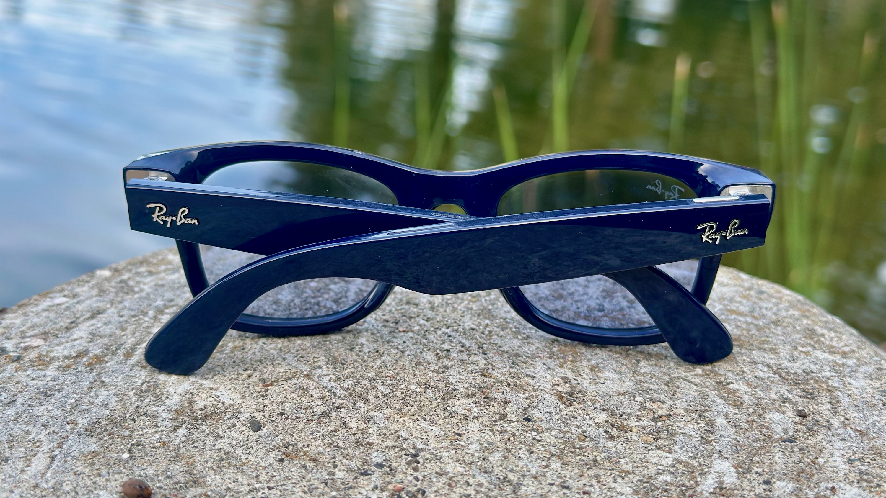 A photo of the Ray-Ban Meta (Gen 2) smart glasses sitting folded up on a rock, angled from behind to show the arms and lenses, with a pond behind them.