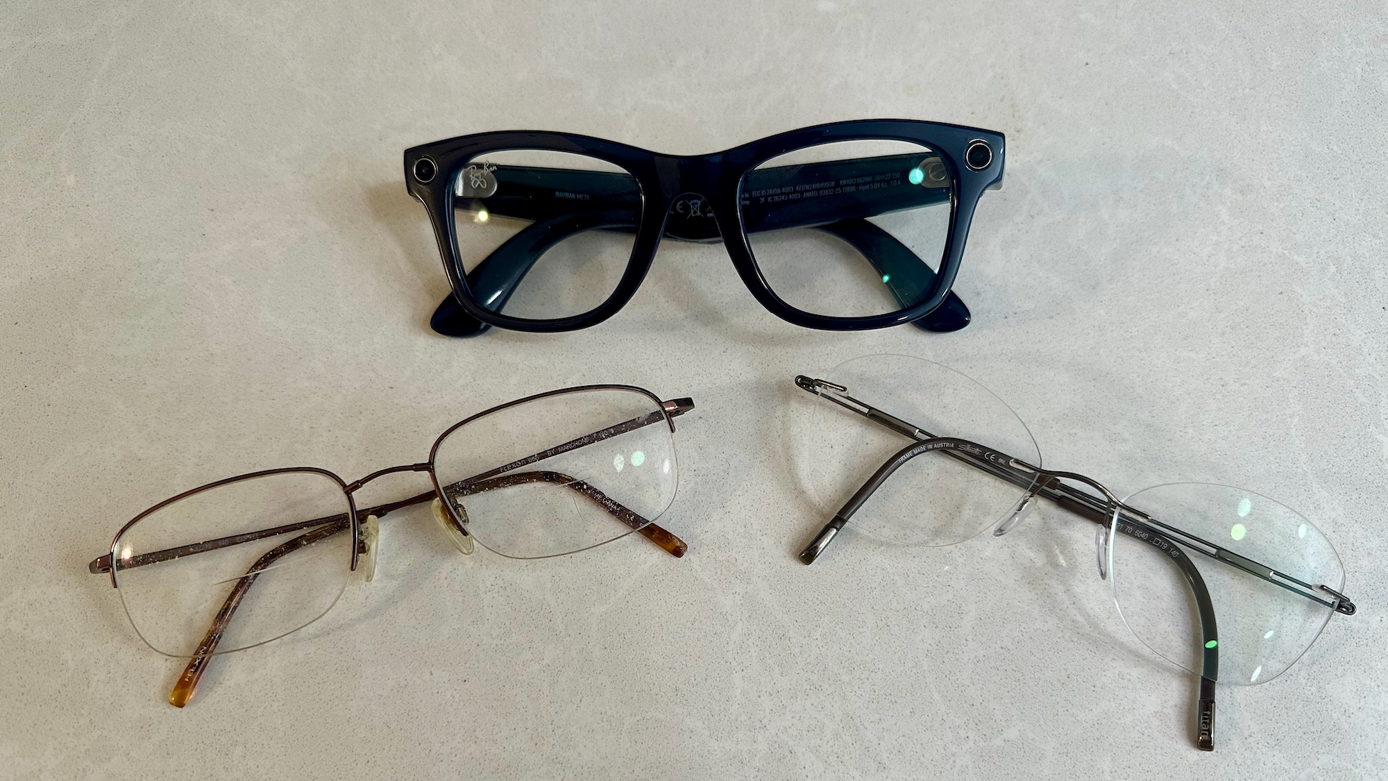 A photo of the Ray-Ban Meta (Gen 2) glasses on a kitchen counter above two pairs of normal prescription glasses with skinnier frames.
