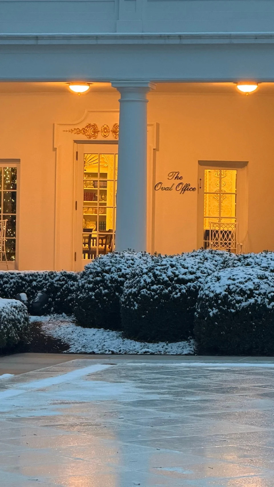 Snow-covered bushes and pathway lead to a warmly lit entrance with a sign reading "The Oval Office." The scene conveys a serene winter setting